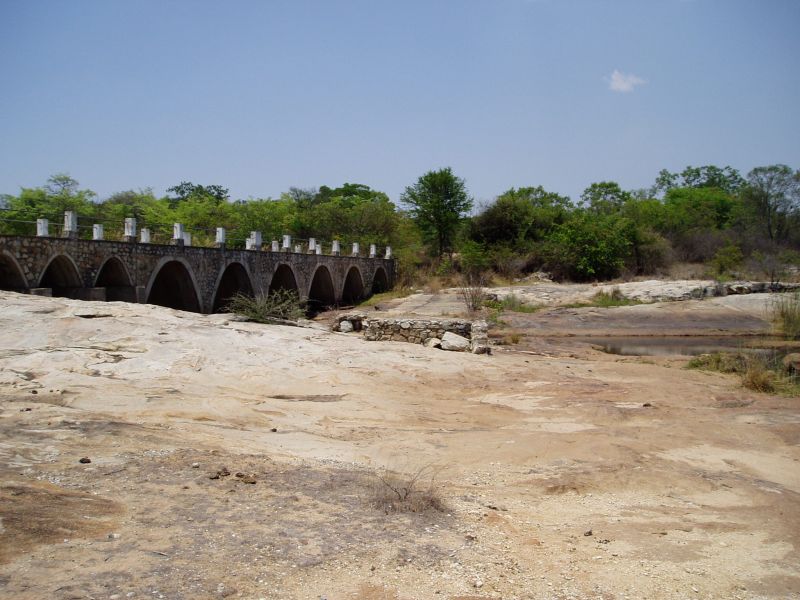 View of bridge from rocky river bed looking southwards towards the south bank. A scarcely flowing water channel may be seen.